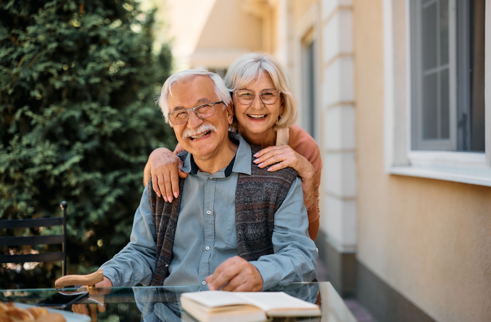 An older adult smiles while hugging their spouse from behind while sitting on an outdoor patio in their new senior living home