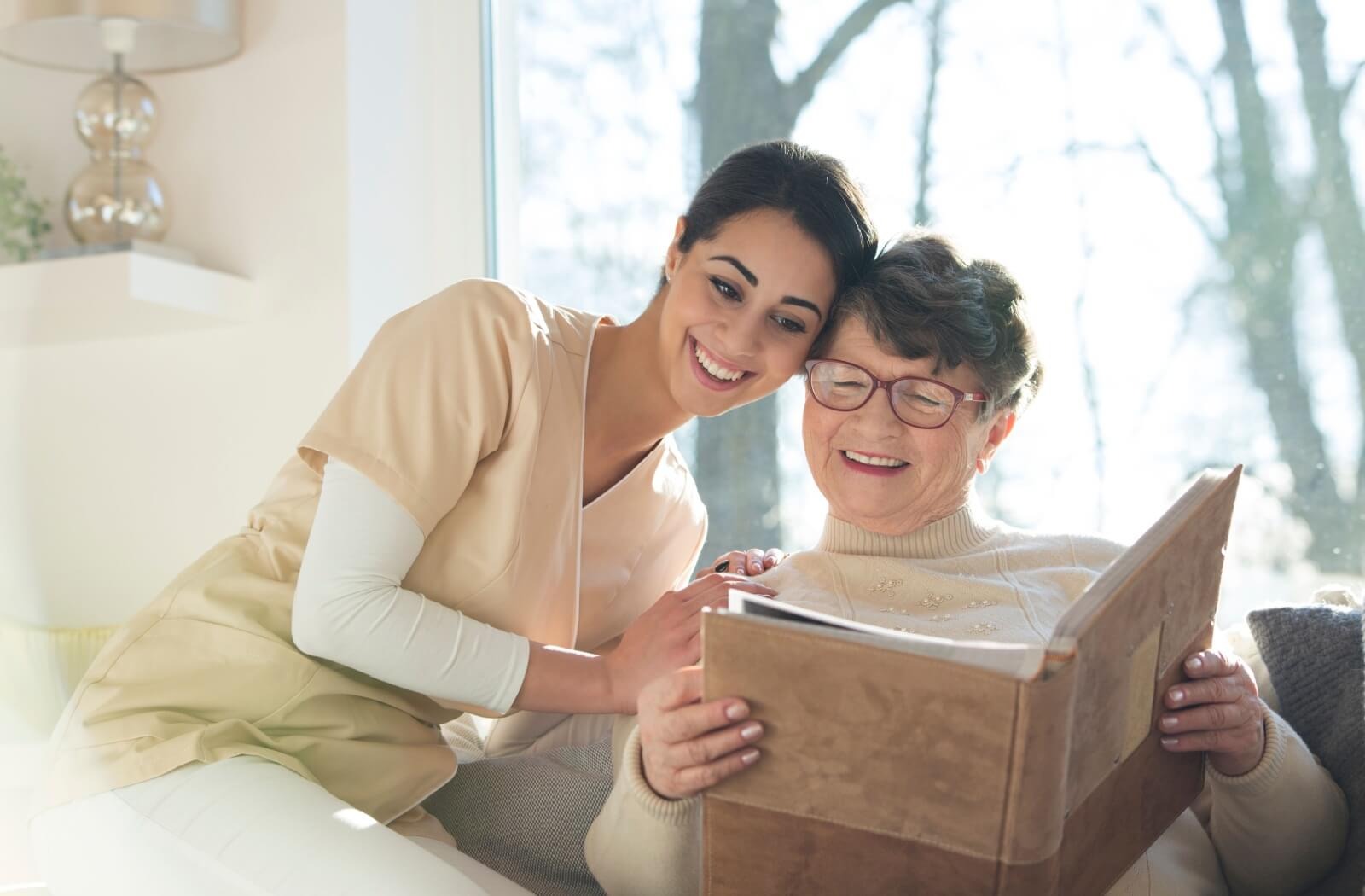 A caregiver touches their head against an older adult's while looking at a photo album together in a sunlit living room.