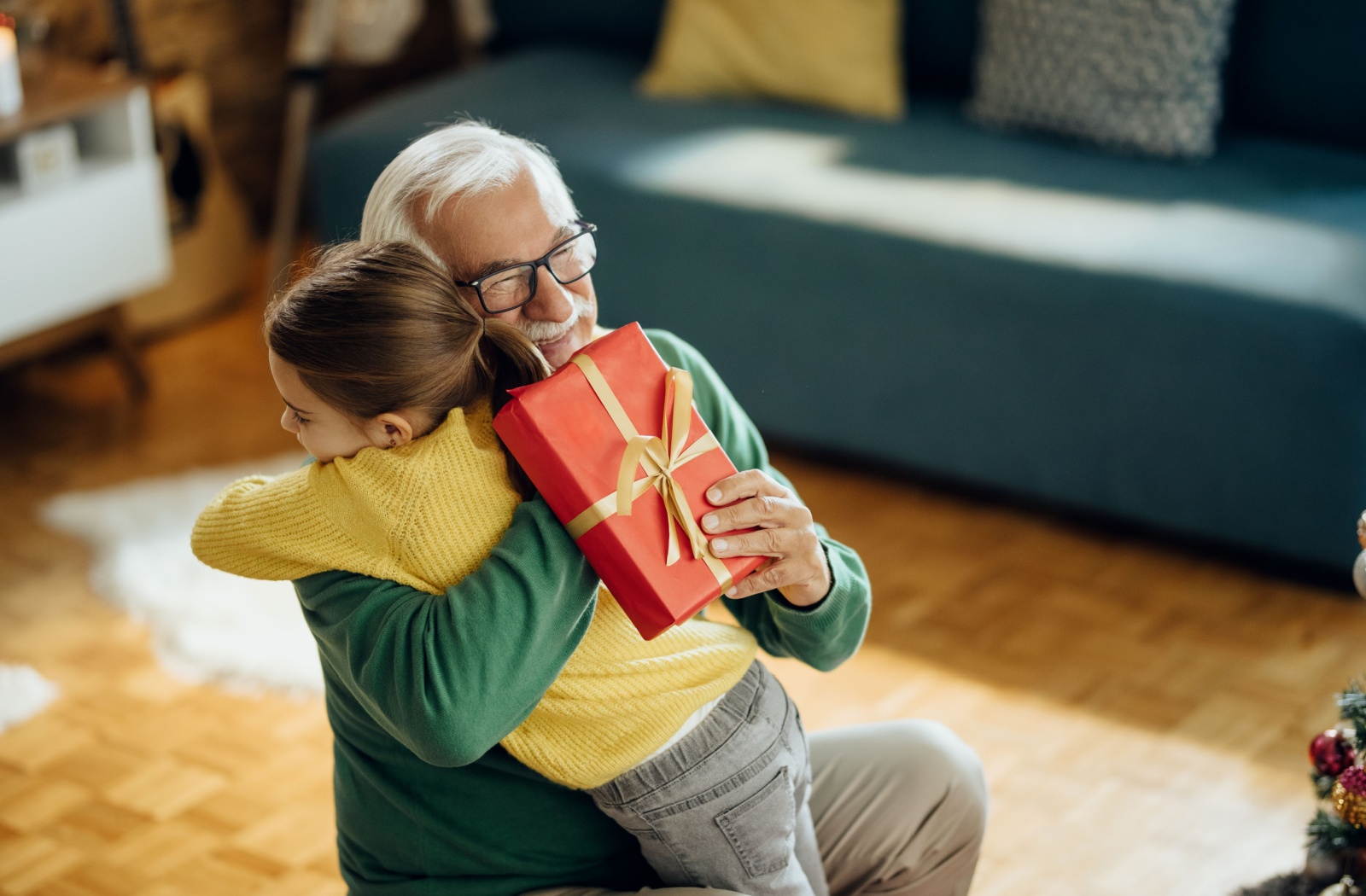 A grandparent crouches down to hug their grandchild while holding a neatly-wrapped gift.
