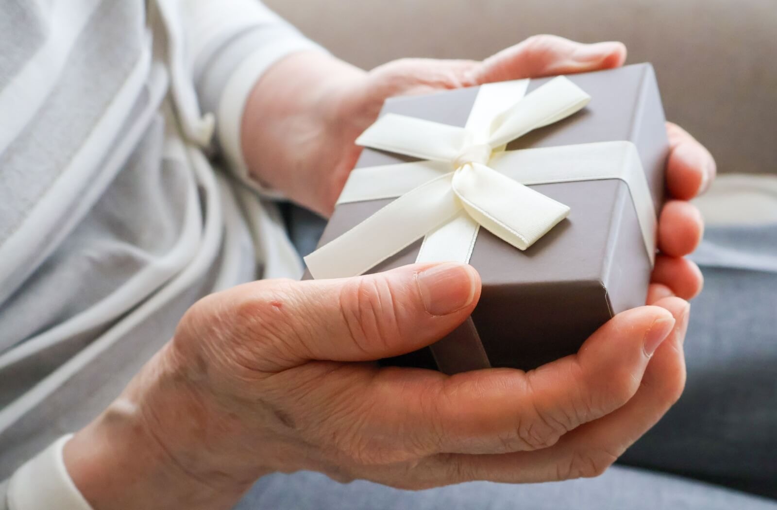 Close up of an older adult holding a neatly-wrapped gray gift box with a white ribbon and bow.
